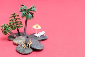 persone in miniatura che indossano costumi da bagno che si rilassano su una roccia con uno sfondo rosa foto