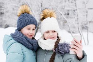 Due sorridente ragazze nel caldo cappotti e cappelli siamo fotografato su un' smartphone nel un' nevoso inverno parco. stile di vita uso di tecnologia foto