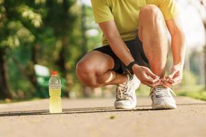 giovane atleta uomo legatura in esecuzione scarpe con energia bevanda acqua, maschio corridore pronto per jogging fuori, asiatico fitness a piedi e esercizio nel il parco mattina. benessere, benessere e sport concetti foto