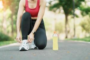 giovane atleta donna legatura in esecuzione scarpe con energia bevanda acqua, femmina corridore pronto per jogging fuori, asiatico fitness a piedi e esercizio nel il parco mattina. benessere, benessere e sport concetti foto