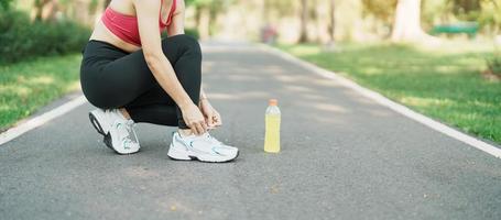 giovane atleta donna legatura in esecuzione scarpe con energia bevanda acqua, femmina corridore pronto per jogging fuori, asiatico fitness a piedi e esercizio nel il parco mattina. benessere, benessere e sport concetti foto