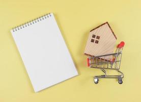 piatto disposizione di di legno Casa modello nel shopping carrello o shopping carrello con vuoto pagina ha aperto taccuino su giallo sfondo, casa Acquista concetto. foto