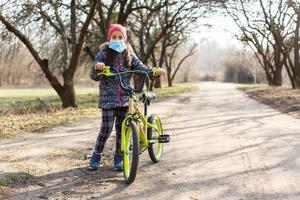 7 anni vecchio contento poco ragazza bambino cavalcata bicicletta nel il parco a casa e indossare protezione maschera per proteggere PM2.5 e coronavirus covid-19 pandemia virus sintomi.sport esercizio per Salute. foto