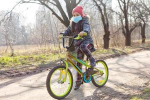 7 anni vecchio contento poco ragazza bambino cavalcata bicicletta nel il parco a casa e indossare protezione maschera per proteggere PM2.5 e coronavirus covid-19 pandemia virus sintomi.sport esercizio per Salute. foto