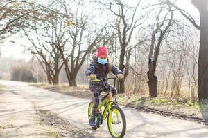 poco ragazza indossare anti inquinamento maschera su sua bicicletta a parco. foto