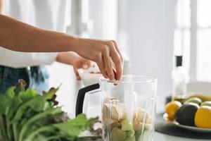 giovane sottile donna nel bianca maglietta e blu jeans cucinando frullato salutare cibo nel cucina a casa foto