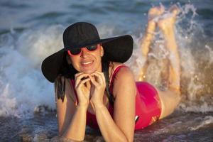 contento anziano donna nel occhiali da sole e un' spiaggia cappello bugie nel il mare onde e sorrisi. foto
