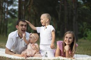 marito e moglie e loro poco bambini. famiglia ritratto nel natura. mamma e papà con loro fratello e sorella siamo dire bugie su il erba. giovane famiglia con bambini per un' camminare. foto