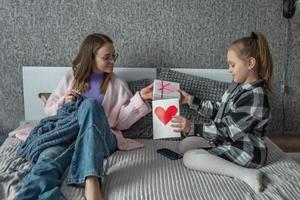 il figlia si congratula sua madre su La madre di giorno, dà sua un' scatola con un' regalo e un' congratulazioni opener con un' cuore fatto di se stessa. un' donna maglia nel sua Camera da letto. foto