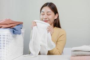 sentire morbidezza, sorridente asiatico giovane donna, ragazza toccante soffice asciugamano cotone, odorare fresco pulito Abiti su tavolo dopo lavaggio, lavanderia, asciutto. domestico Lavorando a casa. lavanderia e domestica. foto