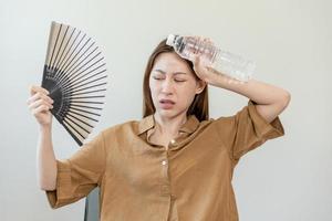 estate calore ictus, caldo tempo atmosferico, stanco asiatico giovane donna sudato e assetato, rinfrescante con mano nel soffiando, onda fan per ventilazione, Tenere freddo acqua bottiglia rubinetto sua corpo quando temperatura alto. foto