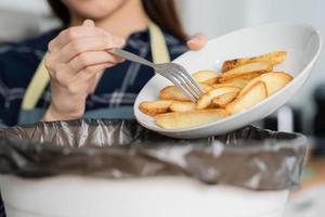 colesterolo di Rifiuto pasto è Grasso pasto, mano di asiatico giovane domestico donna raschiare, lancio cibo avanzi in spazzatura, spazzatura bidone a partire dal Patata patata fritta, merenda. l'ambiente responsabile, ecologia foto