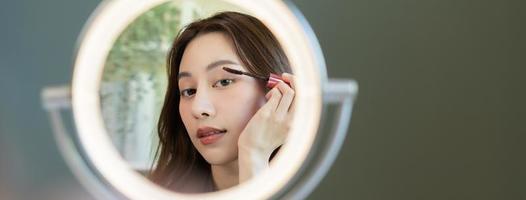 bellezza blogger concetto, carino asiatico giovane donna, ragazza rendere su viso di l'applicazione mascara spazzola su sopracciglio, guardare a il specchio a casa. femmina Guarda con naturale moda stile. foto