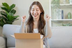 contento emozionato, asiatico giovane donna, ragazza cliente seduta su divano a casa, apertura e disimballaggio cartone scatola scatola di cartone pacco dopo acquisto ordinazione regalo, shopping in linea, consegna servizio concetto. foto