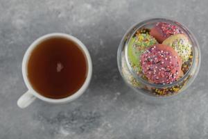 un barattolo di vetro pieno di piccole ciambelle colorate con una tazza di tè caldo foto