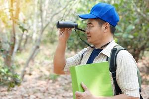 asiatico uomo esploratore indossa blu berretto, detiene binoculare nel foresta per sondaggio botanico impianti e creature animali selvatici. concetto, natura esplorazione. ecologia e ambiente. foto