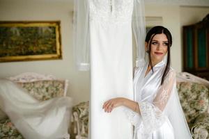 romantico atmosfera di il della sposa mattina. sposa nel un' casa. sposa mattina preparazione. sposa nel bellissimo vestito vicino il indossatrice con vestito in casa a casa. godere ogni momento. foto