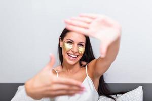 donna con oro cosmetici maschera, occhio cerotti. ritratto di bellissimo giovane donna con oro cosmetici maschera, occhio cerotti. seduta su letto e Spettacoli gesto foto telaio con dita. ritratto