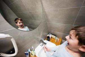 ragazzo lavaggi con divertente viso nel specchio a bagno. foto