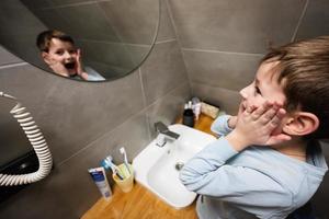 ragazzo lavaggi con divertente viso nel specchio a bagno. foto