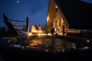 bambini godendo bagnarsi nel di legno barile caldo vasca nel il terrazza di il villetta. scandinavo vasca da bagno con un' camino per bruciare legna e calore acqua. foto