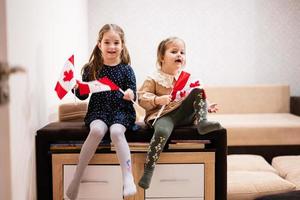 Due sorelle siamo seduta su un' divano a casa con canadese bandiere su mani. Canada bambini ragazze con bandiera . foto