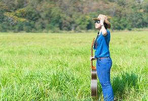 donna indossare cappello e trasportare sua chitarra nel erba campo foto