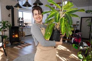 un' contento donna nel un' verde Casa con un' in vaso pianta nel sua mani sorrisi, prende cura di un' fiore. il interno di un' accogliente eco-friendly Casa, un' camino fornello, un' passatempo per in crescita e allevamento homeplant foto