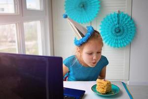 poco ragazza colpi su un' candela su un' torta. festeggiare compleanno attraverso Internet nel quarantena volta, auto-isolamento foto