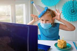 un' contento poco ragazza, storto nel un' medico maschera nel davanti di un' il computer portatile celebra un' compleanno attraverso il Internet nel quarantena volta, auto-isolamento, in linea compleanno. foto