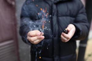 il petardo nel un' mano. ragazzo Tenere un' ardente petardo nel il suo mano. ragazzo con un' pirotecnica quello ustioni con scintille e Fumo all'aperto. petardo e accendino nel mano. foto