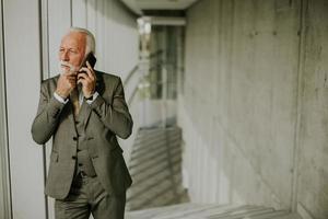 un' anziano attività commerciale uomo sta nel un ufficio corridoio, focalizzata su il suo mobile Telefono foto