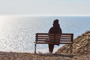 singolo ragazza nel un' nero giacca e cappello seduta su panchina a scogliera a davanti di mare tranquillo, calmo silenzioso posto foto