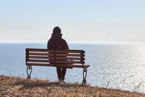 singolo ragazza nel un' nero giacca e cappello seduta su panchina a scogliera a davanti di mare tranquillo, calmo silenzioso posto foto