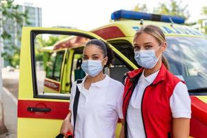 Due femmina paramedici in piedi di il ambulanza. Due emergenza infermieri con viso maschera. paramedico trasporto un' medico trauma Borsa foto