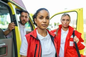 tre multirazziale paramedici in piedi nel davanti di ambulanza veicolo, trasporto portatile attrezzatura foto