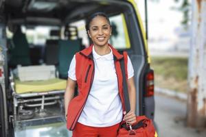 giovane donna , un' paramedico, in piedi a il posteriore di un ambulanza, di il Aperto porte. lei è guardare a il telecamera con un' fiducioso espressione, sorridente, trasporto un' medico trauma Borsa su sua spalla. foto