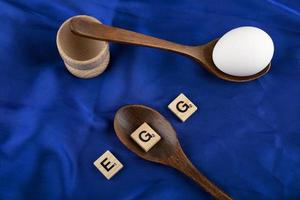 unico uovo crudo con cucchiai di legno e lettere di legno che ortografano la parola uovo su un panno di raso blu foto