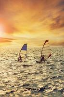 Due uomo giocando vento Surf al di sopra di affrettandosi mare contro bellissimo tramonto illuminazione foto