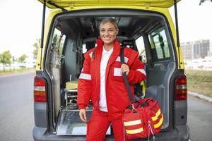 giovane donna , un' paramedico, in piedi a il posteriore di un ambulanza, di il Aperto porte. lei è guardare a il telecamera con un' fiducioso espressione, sorridente, trasporto un' medico trauma Borsa su sua spalla. foto