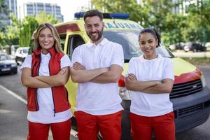 multietnico gruppo di paramedici in piedi a il lato di un ambulanza con Aperto porte. loro collaboratore trasporto un' medico trauma Borsa. essi siamo sorridente a il telecamera. foto