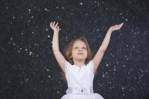 bellissimo poco ragazza nel un' bianca vestito con sollevato braccia sotto volante fiocchi di neve. foto