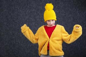 divertente poco ragazzo nel un' soffice giallo pelliccia cappotto e un' a maglia cappello su un' grigio sfondo. foto