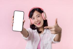 contento allegro sorridente asiatico donna con senza fili auricolari mostrando vuoto schermo mobile Telefono o nuovo smartphone musica applicazione annuncio pubblicitario modello isolato su rosa studio sfondo. foto