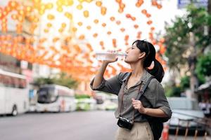 contento giovane asiatico donna zaino viaggiatore potabile un' freddo acqua a Cina cittadina strada cibo mercato nel bangkok, Tailandia. viaggiatore controllo su lato strade. foto