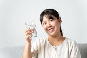 salutare bellissimo giovane donna Tenere un' bicchiere di acqua seduta su il divano a il soggiorno. foto