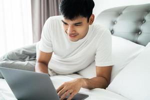 asiatico uomo nel bianca maglietta posa su letto utilizzando il computer portatile computer nel Camera da letto. foto