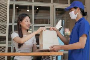 Sorridi, contento asiatico giovane donna o ragazza indossare viso maschera proteggere per pandemia di covid-19 e ricevuto carta Borsa a partire dal consegna uomo, messaggero nel uniforme mentre confinamento, quarantena a davanti porta casa. foto