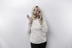 eccitato asiatico donna indossare hijab puntamento a il copia spazio accanto sua mentre Tenere sua Telefono, isolato di bianca sfondo foto