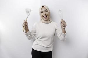 eccitato asiatico musulmano donna indossare un' foulard, Tenere cucinando merce e sorridente, isolato di bianca sfondo foto